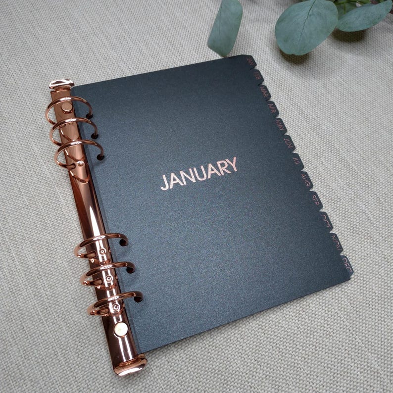 May include: A black planner with a rose gold metal ring binder mechanism on the left side. The cover features the word JANUARY in rose gold foil lettering. The right edge has twelve black index tabs labeled with the months from Jan to Dec.