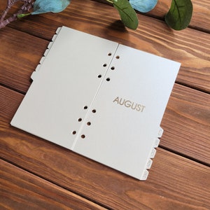 May include: A light gray planner divider page featuring the word AUGUST printed in gold foil. The page includes a series of circular binder holes along the center edge and a set of protruding tabs along the side labeled AUG, SEP, OCT, NOV, and DEC.