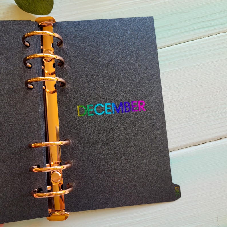 May include: A black planner divider with a metallic copper six-ring binder mechanism on the left side. The word DECEMBER is printed in the center in a vibrant, multi-colored gradient font. A small tab labeled DEC is visible on the bottom right.