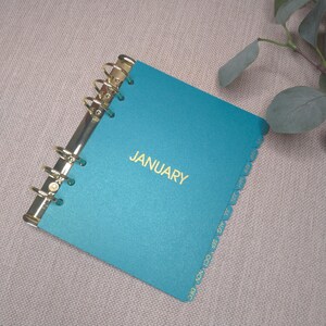 May include: A teal planner with a gold-toned metal ring binding and the word JANUARY printed in gold on the front cover. The planner features side tabs labeled with the months of the year from January through December in gold lettering.