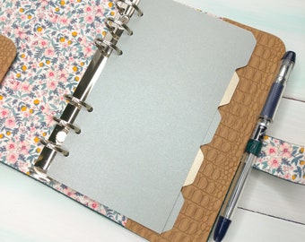May include: A ring-bound planner featuring a floral fabric cover with pink flowers and green leaves, a tan faux-leather panel with a crocodile texture, silver metal rings, a solid silver divider, and a clear plastic pen with a dark blue grip.