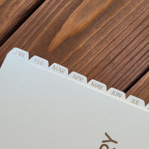 May include: A cream-colored planner or notebook page featuring a series of protruding index tabs along the top edge. Each tab is clearly labeled with the first three letters of the months January through July, printed in a simple, dark sans-serif font.