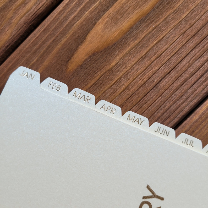 May include: A cream-colored planner or notebook page featuring a series of protruding index tabs along the top edge. Each tab is clearly labeled with the first three letters of the months January through July, printed in a simple, dark sans-serif font.
