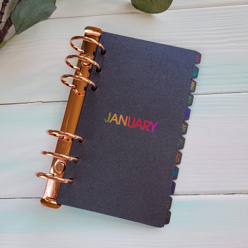 May include: A black personal planner with a metallic rose gold six-ring binder mechanism. The cover features the word JANUARY in a rainbow gradient font. The side includes tabbed dividers labeled with the months of the year in rainbow colors.