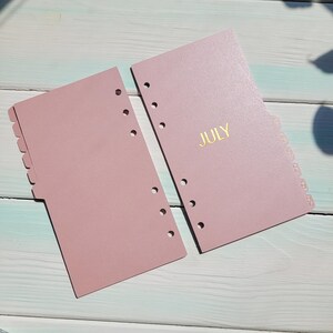 May include: Two dusty rose planner divider sheets with six pre-punched holes along the left side. The right sheet features the word JULY printed in gold foil lettering. Both sheets have a series of tabs along the outer edge for organization.