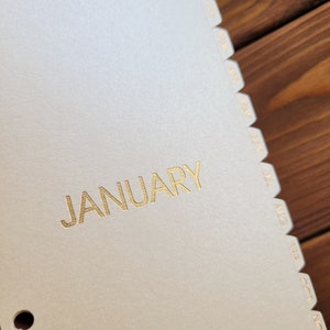 May include: A beige planner divider page with the word JANUARY printed in gold foil lettering. The edge features a series of stepped tabs labeled with the months of the year from February through November in gold foil text, arranged vertically.