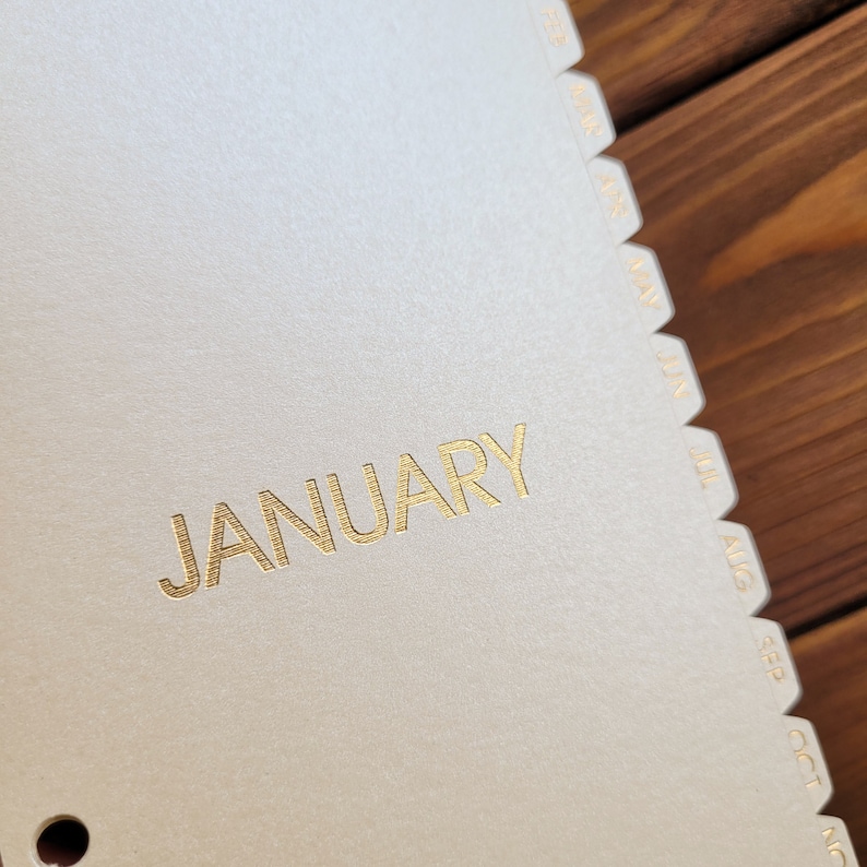 May include: A beige planner divider page with the word JANUARY printed in gold foil lettering. The edge features a series of stepped tabs labeled with the months of the year from February through November in gold foil text, arranged vertically.