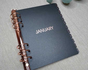 May include: A black planner with a rose gold metal six-ring binder mechanism. The cover features the word JANUARY in rose gold foil lettering. The side edge includes twelve tabbed dividers labeled with the months from JAN to DEC in rose gold.