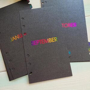 May include: A set of black planner divider pages with punched holes along the left edge. Each page features a month name printed in a vibrant rainbow gradient foil. Visible text includes SEPTEMBER, JANUARY, and TOBER, with small month tabs on the edges.