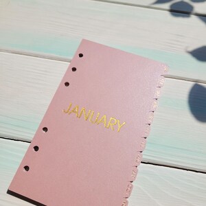 May include: A dusty rose planner divider featuring the word JANUARY printed in gold foil lettering. The rectangular card has six circular punch holes along the left edge and a series of monthly tabs extending from the right side for organization.