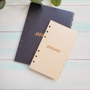 May include: A black planner insert and a beige planner insert, both featuring six punched holes along the left side and side tabs labeled with months from January to December. The beige insert displays the word JANUARY in gold lettering.