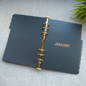 May include: A black planner insert with a gold-toned six-ring metal binder mechanism. The page features the word JANUARY printed in gold foil. The right edge includes a series of monthly tab dividers labeled JAN, FEB, MAR, APR, MAY, JUN, JUL, AUG.