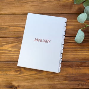 May include: A white planner with the word JANUARY printed in a copper-colored font on the front cover. The planner features a side tab index with abbreviations for each month of the year, from JAN through DEC, arranged vertically along the edge.