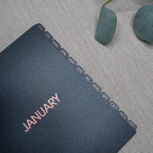 May include: A black planner with rose gold foil lettering that reads JANUARY. The planner features twelve side tabs labeled with the months of the year from JAN to DEC in rose gold foil, providing a clear organizational layout for the entire year.