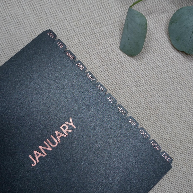 May include: A black planner with rose gold foil lettering that reads JANUARY. The planner features twelve side tabs labeled with the months of the year from JAN to DEC in rose gold foil, providing a clear organizational layout for the entire year.