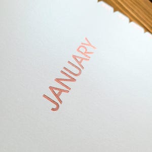 May include: A white paper page with the word JANUARY printed in a metallic rose gold foil. The text features a fine, horizontal line texture within the letters. The top edge of the paper has a scalloped, die-cut design with a metallic finish.