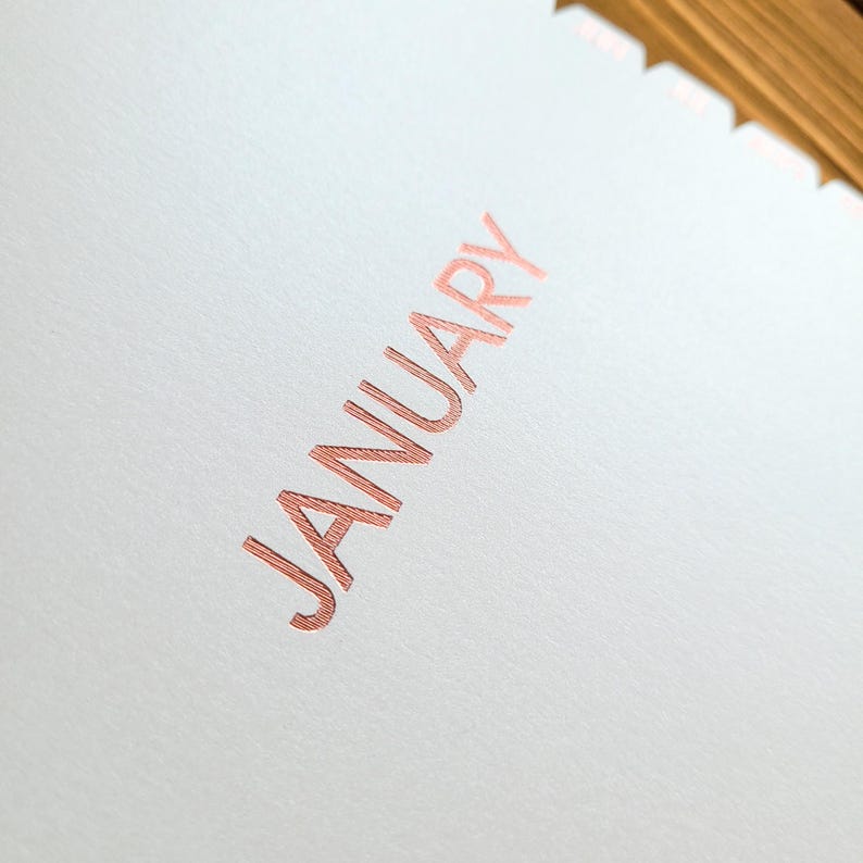 May include: A white paper page with the word JANUARY printed in a metallic rose gold foil. The text features a fine, horizontal line texture within the letters. The top edge of the paper has a scalloped, die-cut design with a metallic finish.