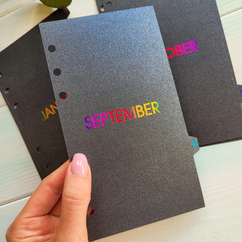 May include: A black planner divider with a textured finish and six punched holes along the left edge. The word SEPTEMBER is printed in a vibrant rainbow gradient foil across the center. Additional black dividers with rainbow text are visible.