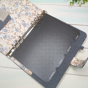 May include: A six-ring binder planner with a denim-textured cover and a floral interior lining featuring blue, brown, and white flowers. The planner includes dark gray tabbed dividers labeled with the text Workouts, Meals, Monthly, and Weekly.
