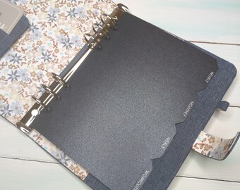 May include: A six-ring binder planner with a denim-textured cover and a floral interior lining featuring blue, brown, and white flowers. The planner includes dark gray tabbed dividers labeled with the text Workouts, Meals, Monthly, and Weekly.