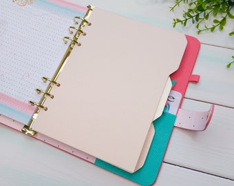 Op de afbeelding: Een roze persoonlijke planner met een gouden zes-rings bandmechanisme, voorzien van een lichtroze tabblad, een turquoise binnenvak en een bijpassende roze riemsluiting. De gestippelde pagina's bevatten een subtiel bloemmotief aan de bovenzijde.
