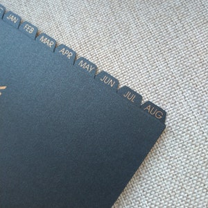 May include: A black index divider sheet featuring gold-colored text for the months of the year. The tabs are labeled JAN, FEB, MAR, APR, MAY, JUN, JUL, and AUG in a clean, sans-serif font. The material has a matte, slightly textured finish.