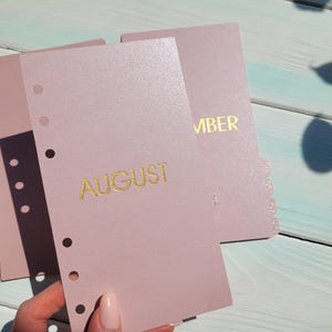 May include: A set of light pink planner dividers with a subtle shimmer finish. Each divider features gold foil lettering for the month, including AUGUST and MBER, and has a vertical row of six circular punched holes along the left side.