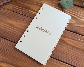 May include: A cream-colored planner divider page with six circular holes along the left edge. The word JANUARY is printed in gold in the center. The right edge features twelve cascading tabs labeled with the months from JAN to DEC in gold text.