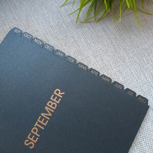 May include: A black planner or notebook featuring gold-colored text that reads SEPTEMBER. The page edges include twelve protruding index tabs, each labeled with a month abbreviation from SEP through AUG in gold lettering against a black background.