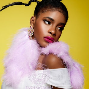 May include: A woman with a braided hairstyle wears a white off-the-shoulder top and a lavender feather boa. She is wearing large, colorful crystal earrings. The background is a bright yellow.
