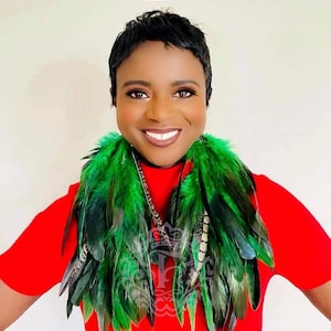 May include: A woman with short black hair is wearing a red shirt and a green feather boa. The boa is made of long, flowing feathers that are arranged in a cascade around her neck.