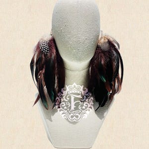 Może przedstawiać: Para kolczyków z piórami, prezentowanych na głowie manekina. Kolczyki mają mieszankę ciemnobrązowych, czarnych i zielonych piór, z kilkoma nakrapianymi białymi i czarnymi piórami. Są przymocowane do srebrnego metalowego haczyka.