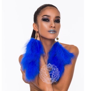 Op de afbeelding: Een paar lange, blauwe veren oorbellen met gouden accenten. De oorbellen worden gedragen door een persoon met donker haar en make-up.