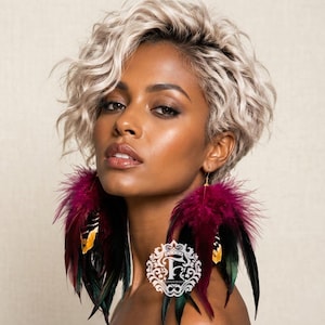 May include: A woman with short, wavy blonde hair is wearing large feather earrings. The earrings feature a cluster of burgundy feathers at the top, transitioning to black and green feathers with yellow accents. The earrings are suspended from gold-colored hooks.
