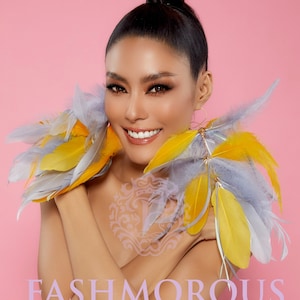 May include: A woman with long black hair pulled back in a bun is wearing large feather earrings. The earrings are made of gray and yellow feathers. The woman is smiling and looking at the camera. The background is a light pink. The text "FASHMOROUS Unique Glamorous Craft Jewelry" is at the bottom of the image.