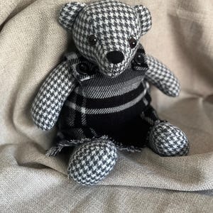 Op de afbeelding: Een knuffelbeer gemaakt van grijs en wit pied-de-poule-stof. De beer draagt een zwart, wit en grijs geruit kledingstuk in tuinbroekstijl met twee kleine zwarte knoopjes op de schouderbanden, een zwarte neus en donkere ogen.