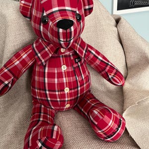 Op de afbeelding: Een knuffelbeer gemaakt van een rood, wit en zwart geruit overhemd. De beer heeft een zwarte neus, donkere ogen en een kraag. Op de linkervoet is de naam James Burdie Bar geborduurd en op de rechtervoet 1962-2021.
