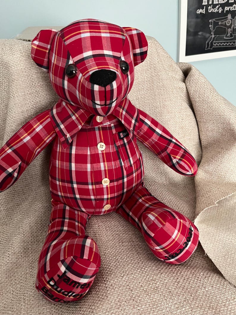 Op de afbeelding: Een knuffelbeer gemaakt van een rood, wit en zwart geruit overhemd. De beer heeft een zwarte neus, donkere ogen en een kraag. Op de linkervoet is de naam James Burdie Bar geborduurd en op de rechtervoet 1962-2021.