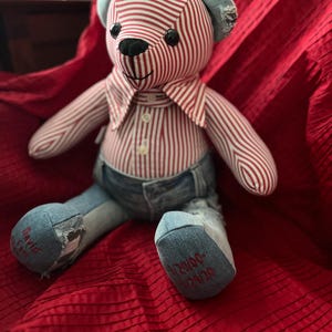 Op de afbeelding: Een knuffelbeer gemaakt van een rood-wit gestreept overhemd en blauwe spijkerstof. De beer heeft zwarte knoopogen, een zwarte geborduurde neus en een kraag. De linkervoet is geborduurd met David en de rechtervoet met 9/20/00-1/29/20.