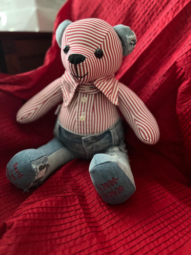Op de afbeelding: Een knuffelbeer gemaakt van een rood-wit gestreept overhemd en blauwe spijkerstof. De beer heeft zwarte knoopogen, een zwarte geborduurde neus en een kraag. De linkervoet is geborduurd met David en de rechtervoet met 9/20/00-1/29/20.
