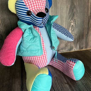Op de afbeelding: Een patchwork teddybeer gemaakt van verschillende stoffen met patronen zoals roze-witte vichyruitjes, blauw-witte strepen en blauwe stippen. De beer draagt een licht turquoise gewatteerd vest met een rits aan de voorzijde en heeft donkere ogen.