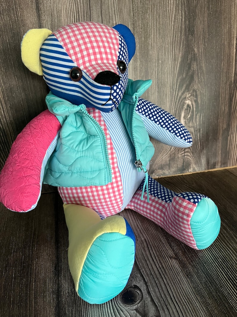 Op de afbeelding: Een patchwork teddybeer gemaakt van verschillende stoffen met patronen zoals roze-witte vichyruitjes, blauw-witte strepen en blauwe stippen. De beer draagt een licht turquoise gewatteerd vest met een rits aan de voorzijde en heeft donkere ogen.
