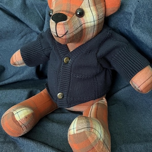 Op de afbeelding: Een knuffelbeer gemaakt van oranje, bruine en cr&egrave;mekleurige ruitjesstof. De beer heeft zwarte plastic ogen, een geborduurde zwarte neus en mond, en draagt een donker marineblauw gebreid vest met twee kleine bruine knoopjes.