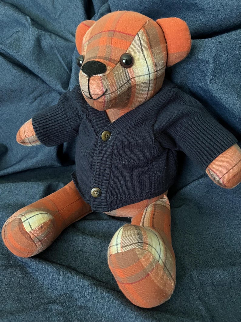 Op de afbeelding: Een knuffelbeer gemaakt van oranje, bruine en cr&egrave;mekleurige ruitjesstof. De beer heeft zwarte plastic ogen, een geborduurde zwarte neus en mond, en draagt een donker marineblauw gebreid vest met twee kleine bruine knoopjes.