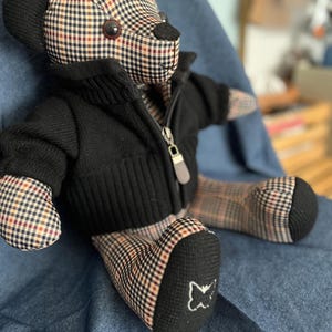 Op de afbeelding: Een knuffelbeer met een beige, zwart en rood ruitpatroon op het lichaam, het hoofd en de ledematen. De beer draagt een zwart gebreid vest met rits en een zilverkleurige ritstrekker. Een klein wit geborduurd vlinderlogo staat op de rechtervoet.