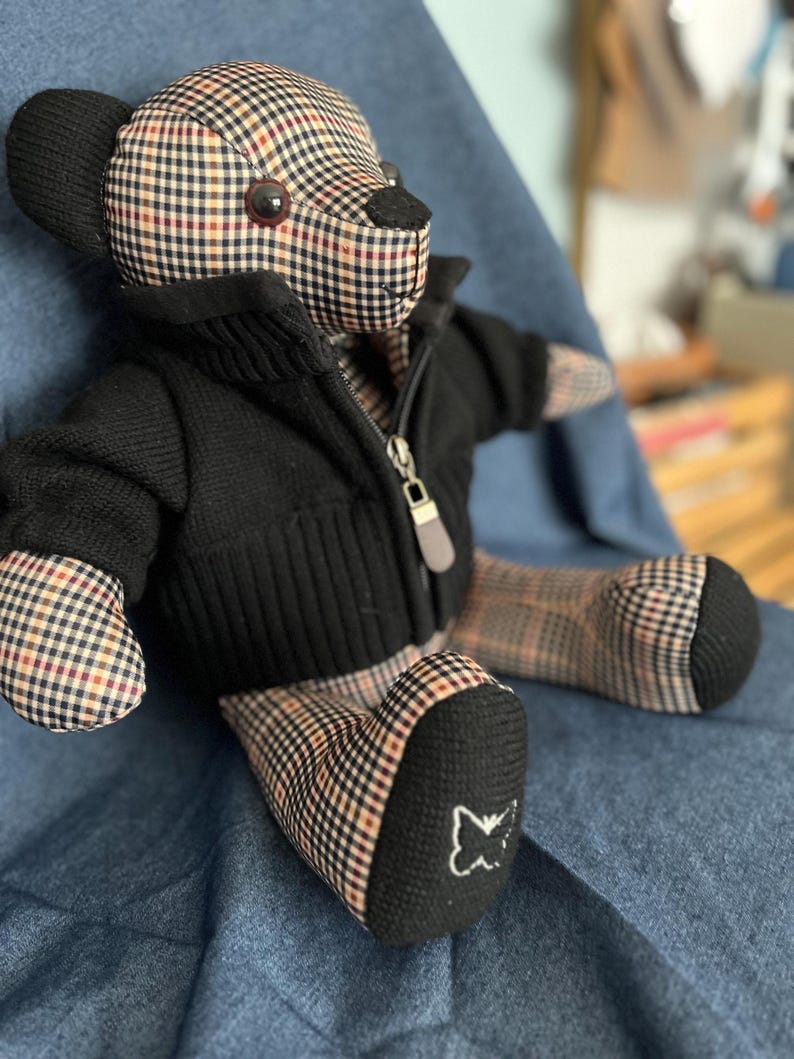 Op de afbeelding: Een knuffelbeer met een beige, zwart en rood ruitpatroon op het lichaam, het hoofd en de ledematen. De beer draagt een zwart gebreid vest met rits en een zilverkleurige ritstrekker. Een klein wit geborduurd vlinderlogo staat op de rechtervoet.