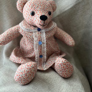 Op de afbeelding: Een knuffelbeer gemaakt van stof met een klein rood en blauw bloemenpatroon. De beer draagt een bijpassend jurkje met een witte kanten rand langs de kraag en de sluiting, vastgezet met drie kleine blauwe knoopjes en een zwarte neus.