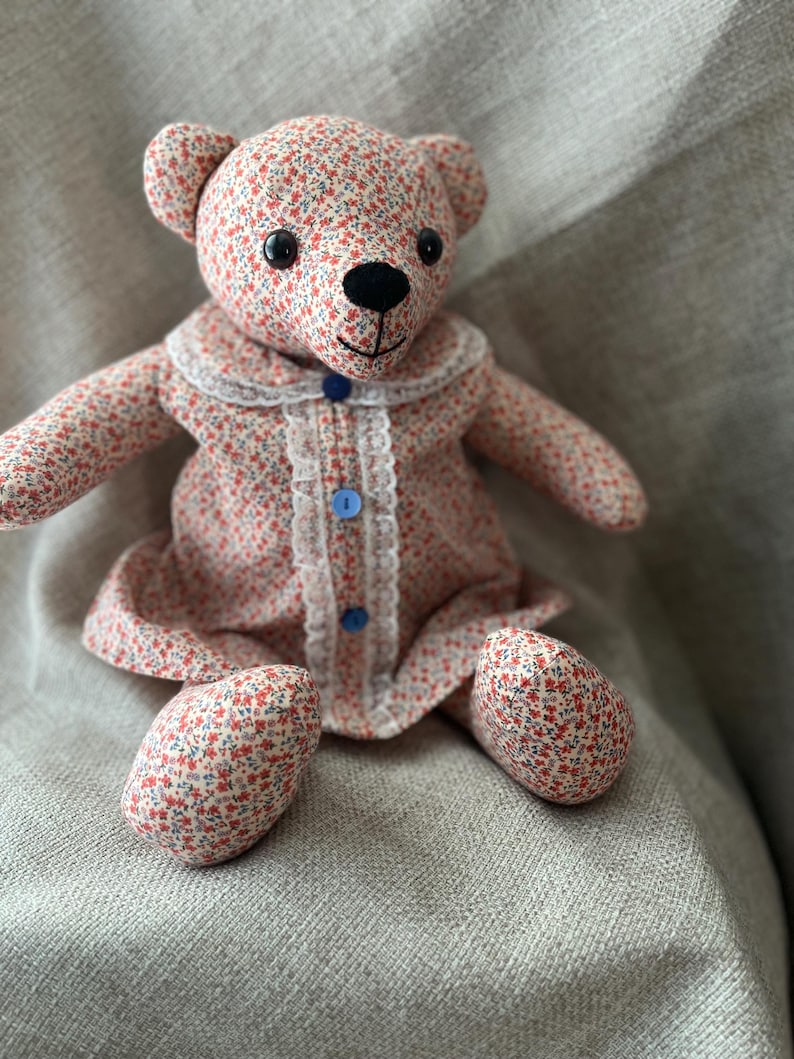 Op de afbeelding: Een knuffelbeer gemaakt van stof met een klein rood en blauw bloemenpatroon. De beer draagt een bijpassend jurkje met een witte kanten rand langs de kraag en de sluiting, vastgezet met drie kleine blauwe knoopjes en een zwarte neus.