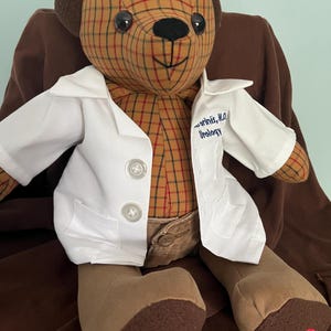 Op de afbeelding: Een knuffelbeer met een hoofd en romp in een bruin en beige ruitpatroon, bruine ledematen en donkerbruine oren. De beer draagt een witte doktersjas met twee knopen en de tekst "[Name], M.D. Urology" op de borst, en een beige broek met een knoop.