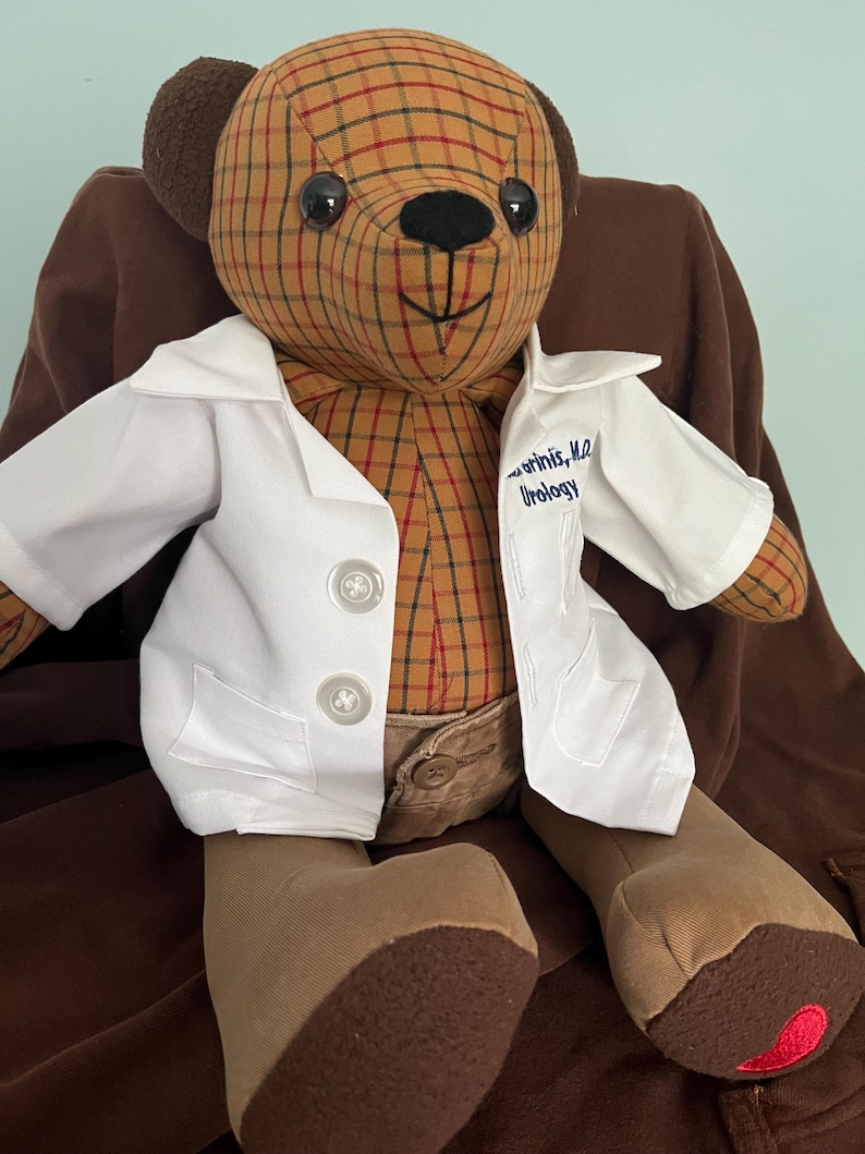 Op de afbeelding: Een knuffelbeer met een hoofd en romp in een bruin en beige ruitpatroon, bruine ledematen en donkerbruine oren. De beer draagt een witte doktersjas met twee knopen en de tekst "[Name], M.D. Urology" op de borst, en een beige broek met een knoop.